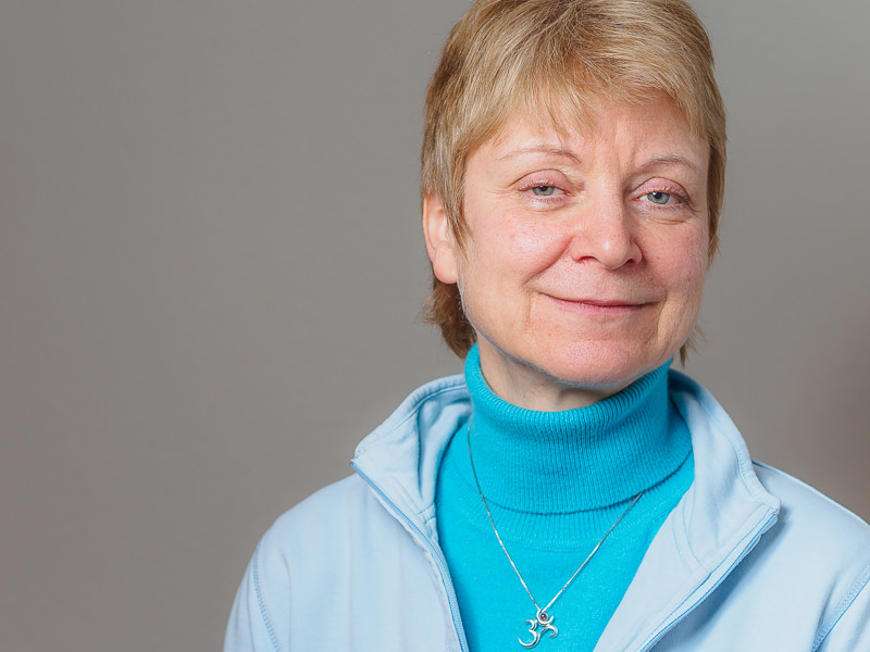 Eine Frau mit blondem Haar trägt einen blauen Rollkragenpullover und eine hellblaue Jacke, sie lächelt sanft in die Kamera.